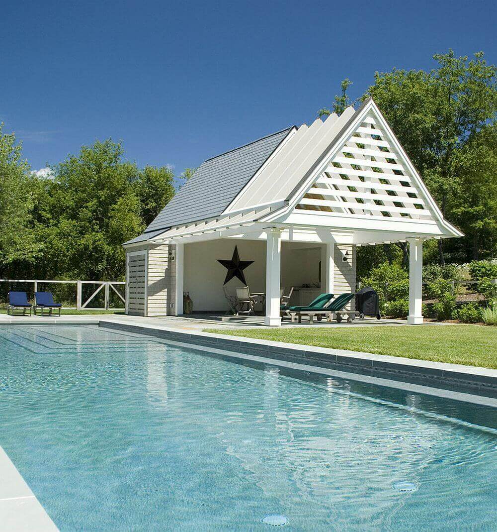 A backyard pool house with a gabled roof featuring white slatted details, adjacent to a rectangular in-ground pool, surrounded by lush greenery.