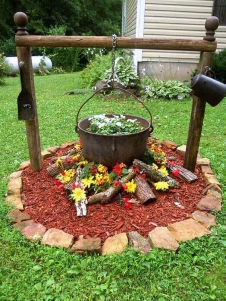 A whimsical flower bed surrounds a wooden garden well, topped with a metal pot, filled with faux flowers and red mulch, enhancing curb appeal.