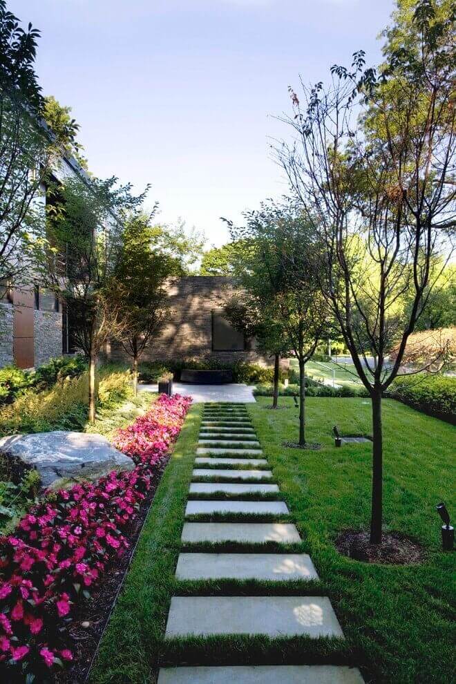Narrow stone pathway flanked by vibrant pink begonias, leading through ornamental grasses and trees, enhancing modern landscape design.