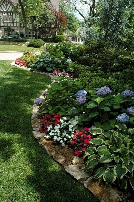 Curvilinear flower bed with hydrangeas, red begonias, and white impatiens, framed by a 6-inch stone border, enhancing curb appeal.