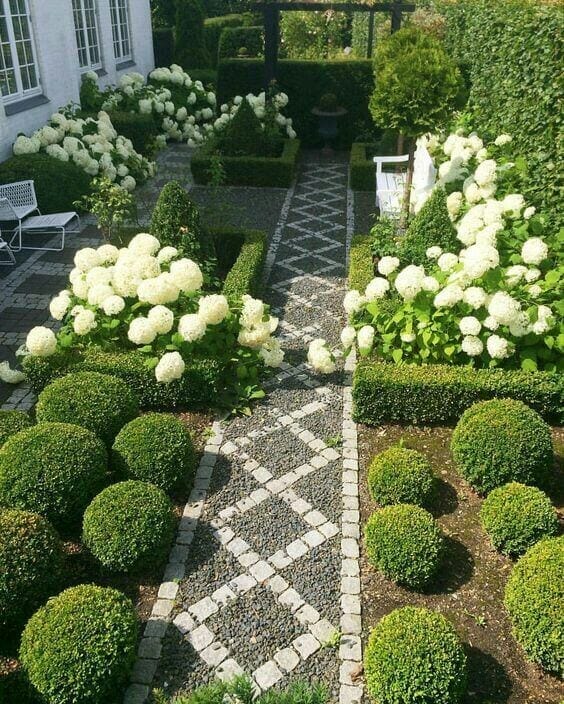 Structured garden layout with a central cobblestone path, flanked by geometric boxwood hedges and clusters of white hydrangeas.