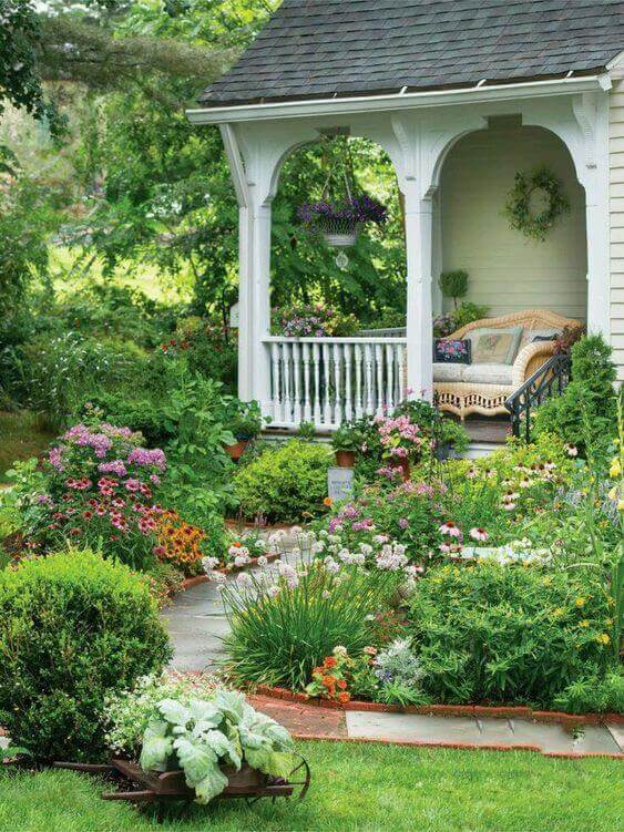 Cottage-style garden with bright flowering plants and lush greenery around a tiled pathway, leading to a shaded porch.