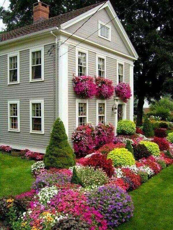 Charming two-story house with a grey clapboard exterior, surrounded by vibrant flowerbeds of petunias, begonias, and marigolds.