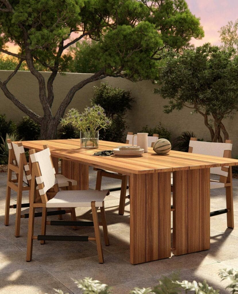 A wooden outdoor dining table set against a serene garden backdrop with plush chairs, a vase of fresh plants at the center, and elegant table settings, all bathed in soft, natural light.