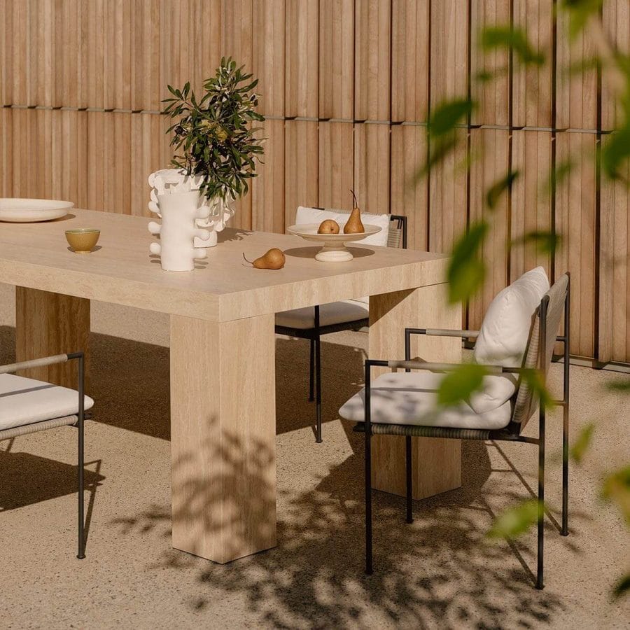 Contemporary outdoor dining setup featuring a minimalist beige stone table adorned with fresh pears, a unique vase holding greenery, and sleek metal-framed chairs, all set against a wooden slatted backdrop with soft shadows from nearby plants.