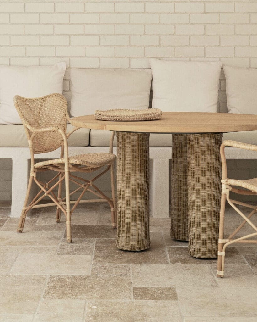 Cozy outdoor dining area showcasing a wooden table with woven rattan pedestal base, paired with natural wicker chairs, set against a white brick wall with cushioned seating and a neutral stone floor.