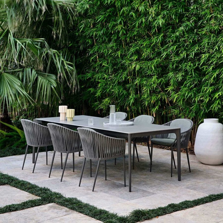 Modern outdoor dining setup with a sleek gray table, surrounded by ribbed-back chairs, set against a lush backdrop of tall bamboo and tropical foliage, complemented by a large white ceramic vase and minimalist table decor.