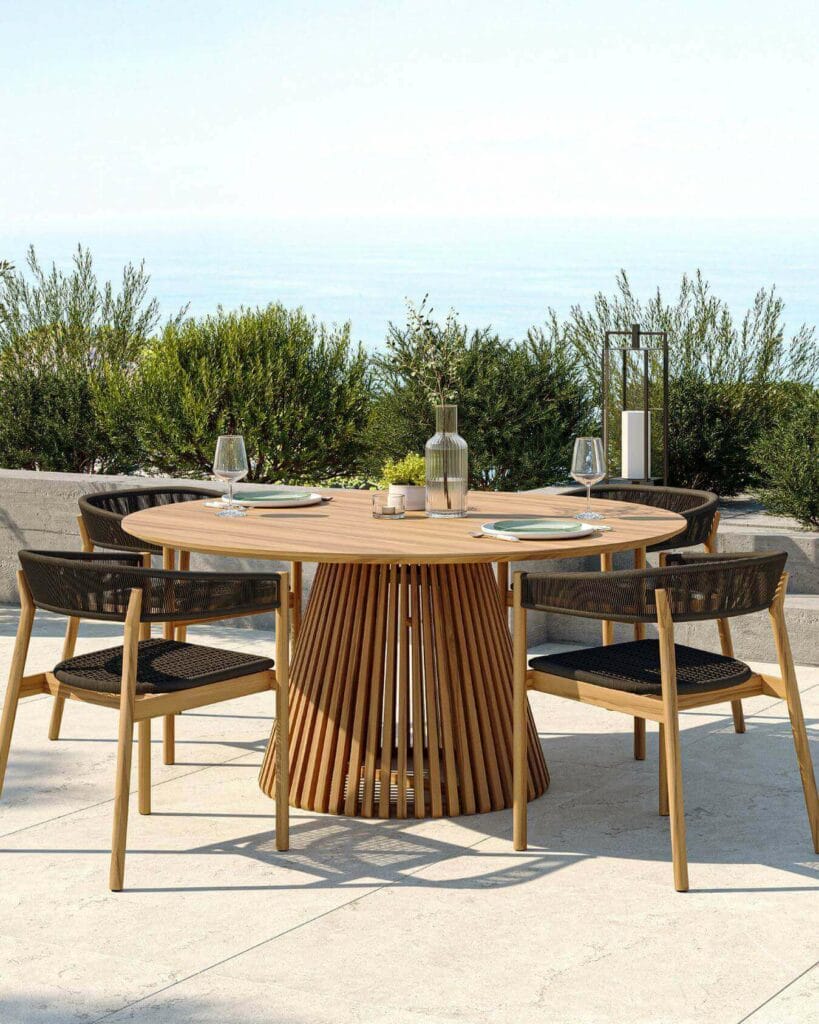 Elegant round wooden outdoor dining table with a unique slatted base, accompanied by woven-back chairs, set against a serene backdrop of shrubs and a distant sea view, adorned with glassware, dishes, and decorative lanterns.
