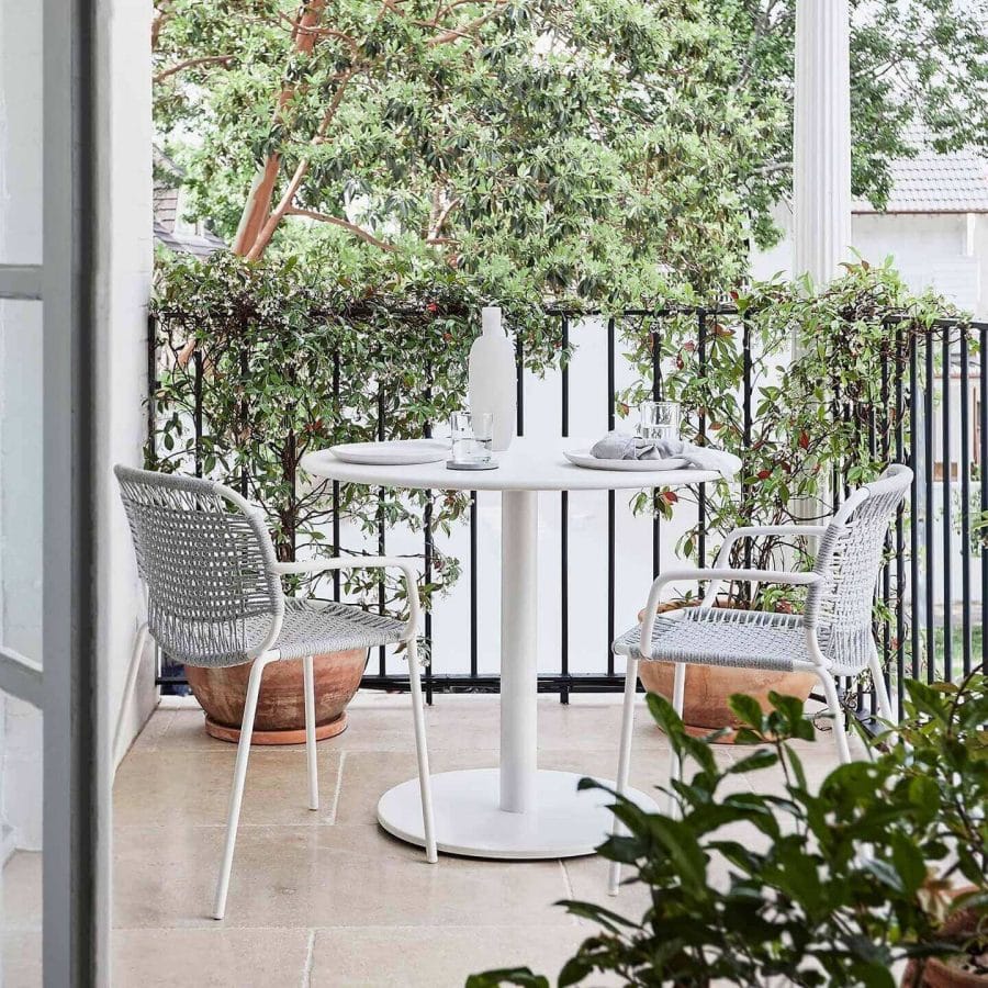 Contemporary white outdoor dining table with a sleek pedestal base, paired with intricately woven gray chairs, set on a balcony overlooking lush greenery, with table settings including a bottle and clear glassware.