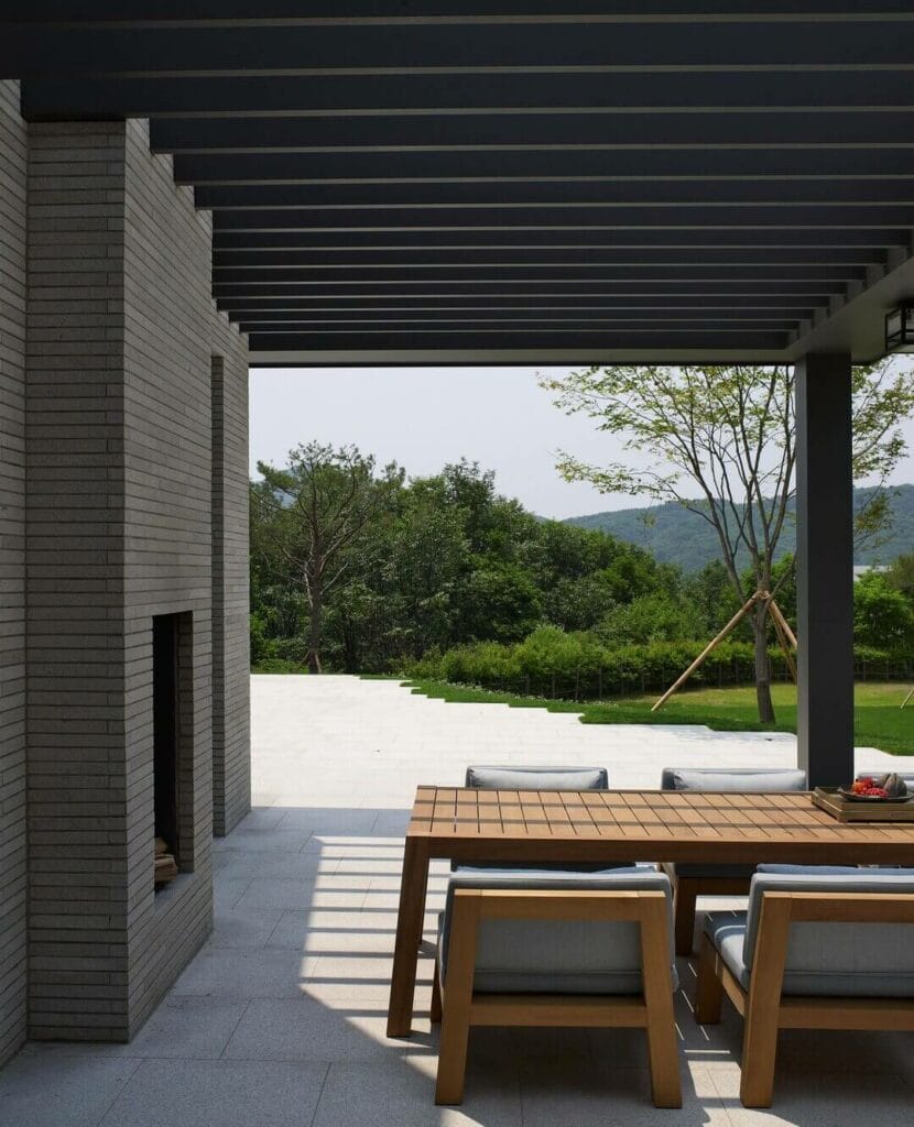 Modern outdoor dining setup under a sleek black pergola next to a brick facade, featuring a wooden table with plush seating, overlooking a manicured lawn with distant trees and hills.