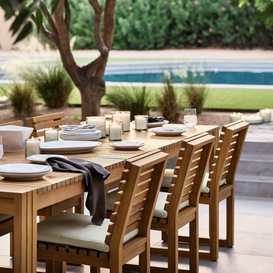 Inviting outdoor dining area with a spacious wooden table set with white dishes, candles, and draped napkins, surrounded by cushioned chairs, positioned near a serene pool and shaded by a mature tree.