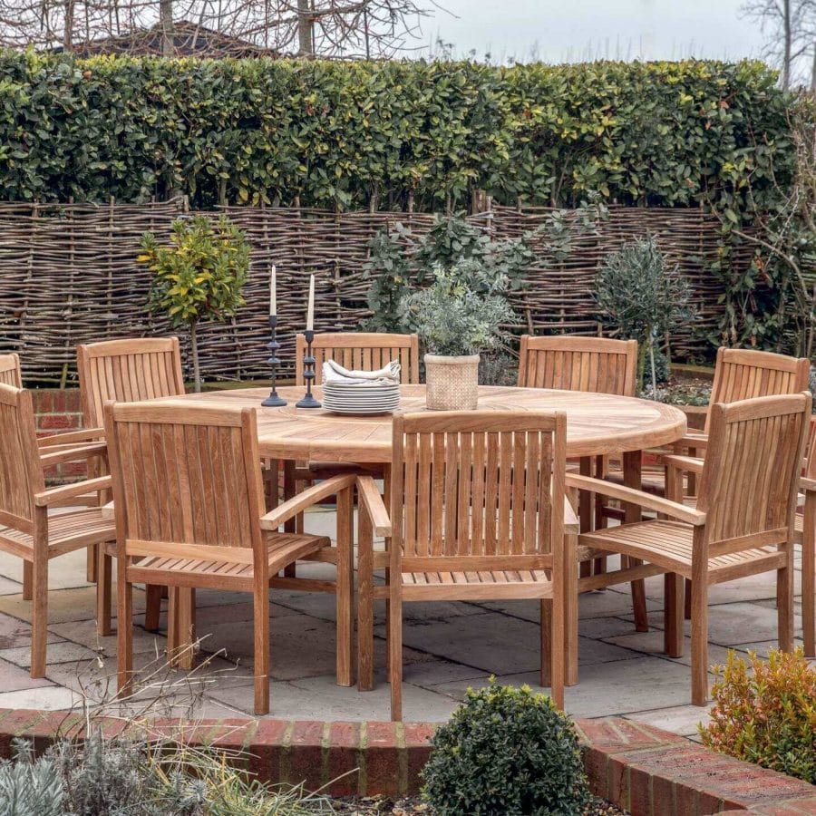 Traditional round wooden outdoor dining table set with matching chairs, adorned with a candlestick and dishes, situated on a paved patio with a woven fence and dense foliage backdrop.