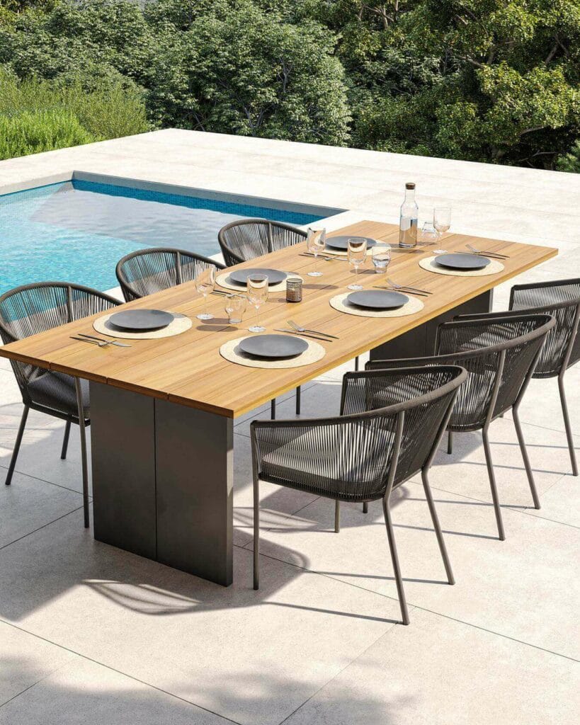 A sleek outdoor dining table with a wooden top and modern metal base, accompanied by stylish wicker chairs, set beside a tranquil swimming pool. The table is elegantly laid with plates, glasses, and silverware, ready for a sophisticated alfresco dining experience amidst lush greenery.