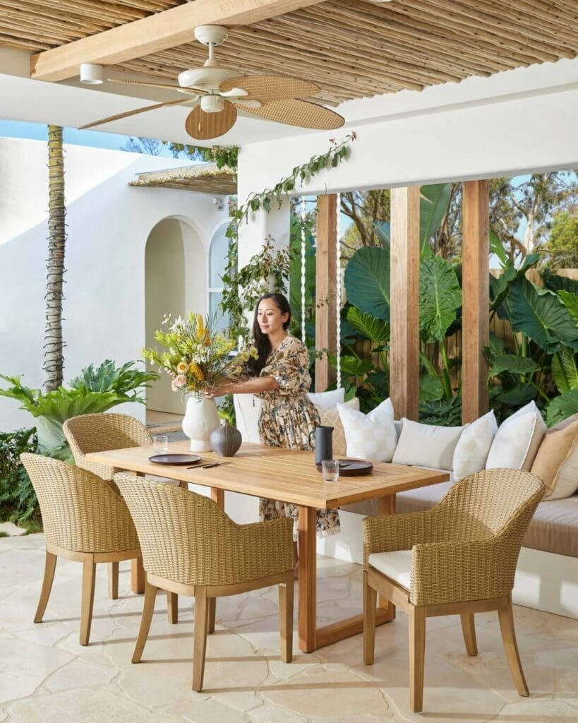 Elegant outdoor dining area under a wooden pergola, featuring a wooden table with woven chairs. A woman in a floral dress arranges a vase of fresh flowers on the table, with a backdrop of lush tropical plants and a white archway.