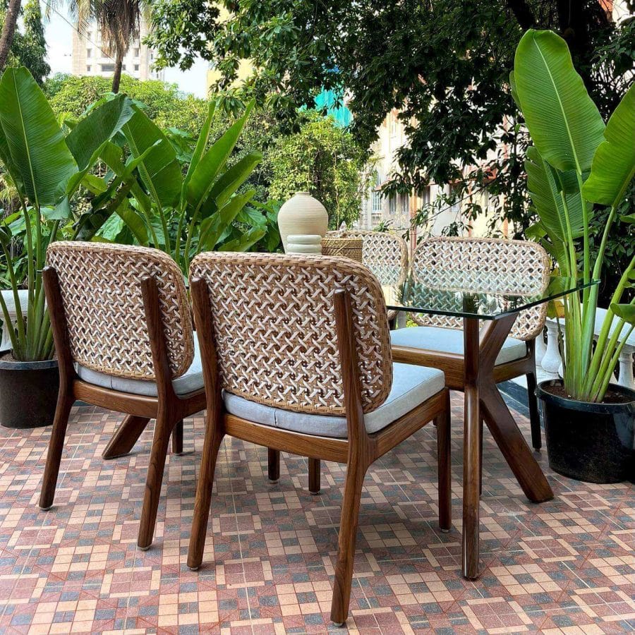 Chic outdoor dining setup with intricately woven back chairs and a glass-top table. Set against a backdrop of urban buildings and surrounded by lush tropical greenery and potted plants on a mosaic-tiled floor.