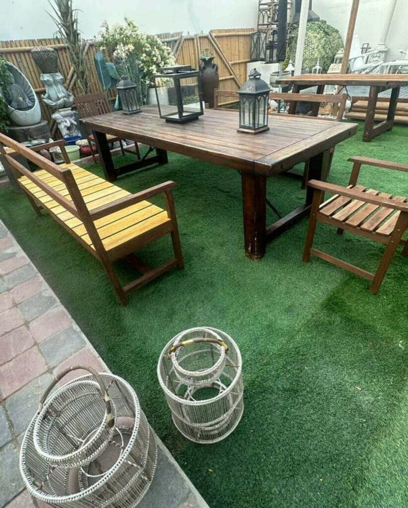 Rustic outdoor dining area featuring a wooden table and benches on a vibrant green artificial grass, complemented by decorative lanterns, birdcages, and assorted garden ornaments, with a bamboo fence backdrop.
