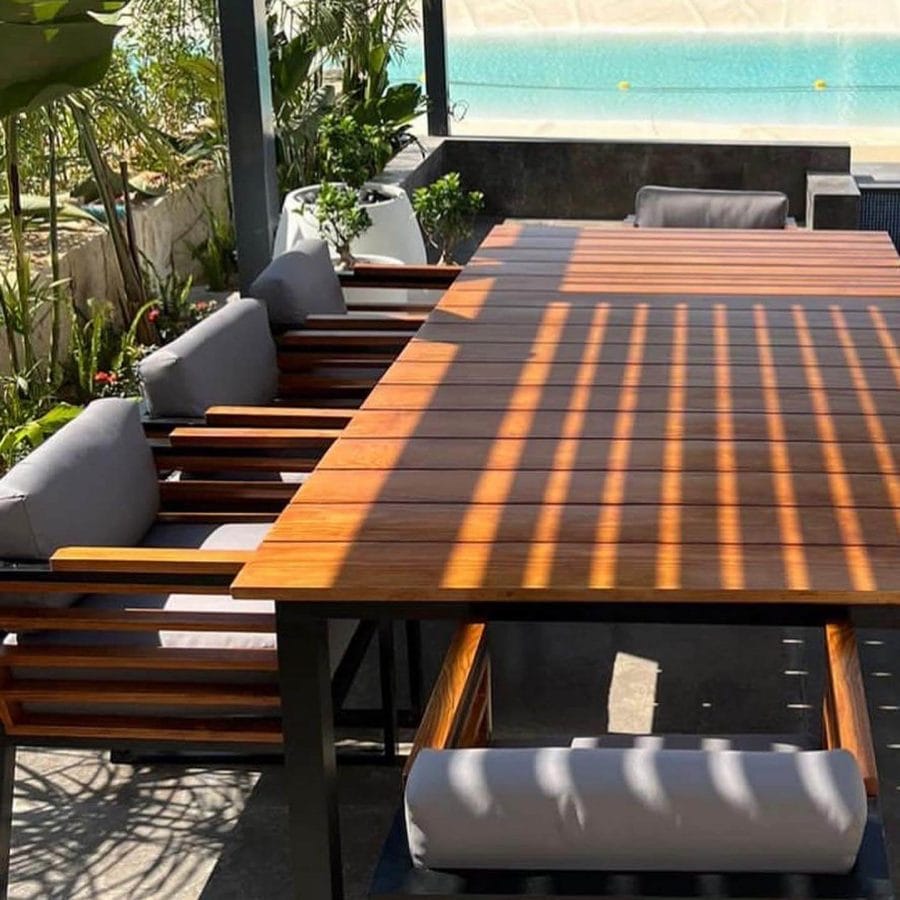 Sleek outdoor dining table with a polished wooden surface displaying distinctive stripe patterns from sunlight filtering through overhead beams, accompanied by cushioned benches, set against a tranquil pool backdrop.