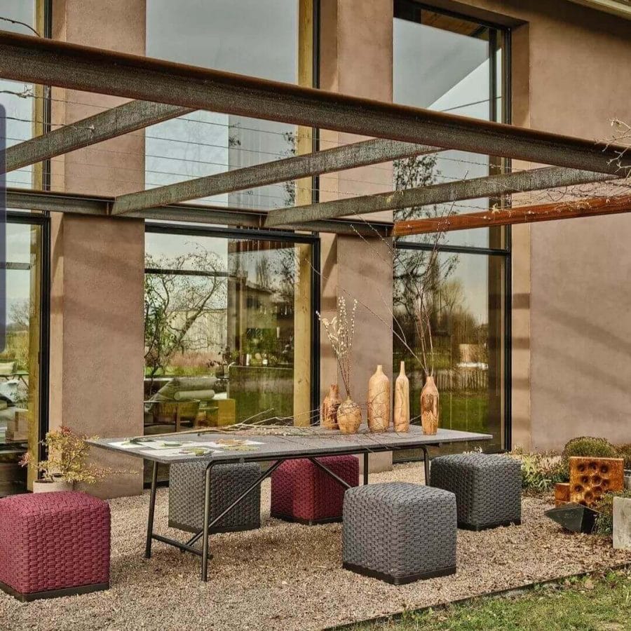 Modern outdoor dining table with a textured surface, adorned with an assortment of artistic ceramic vases, complemented by woven cube stools in shades of red and gray, set against the backdrop of a contemporary home with expansive glass windows.