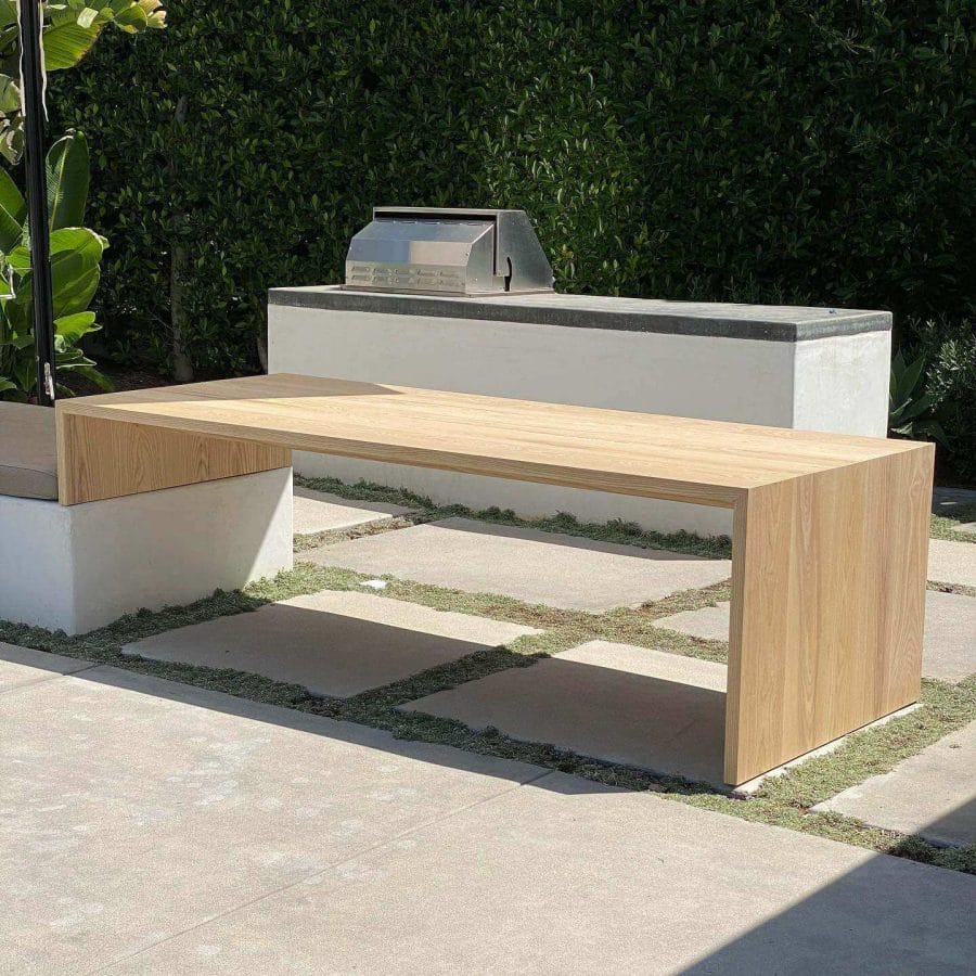 Minimalist outdoor dining table made of smooth light-toned wood, featuring a block design, positioned near a built-in grill against a backdrop of a lush green hedge.