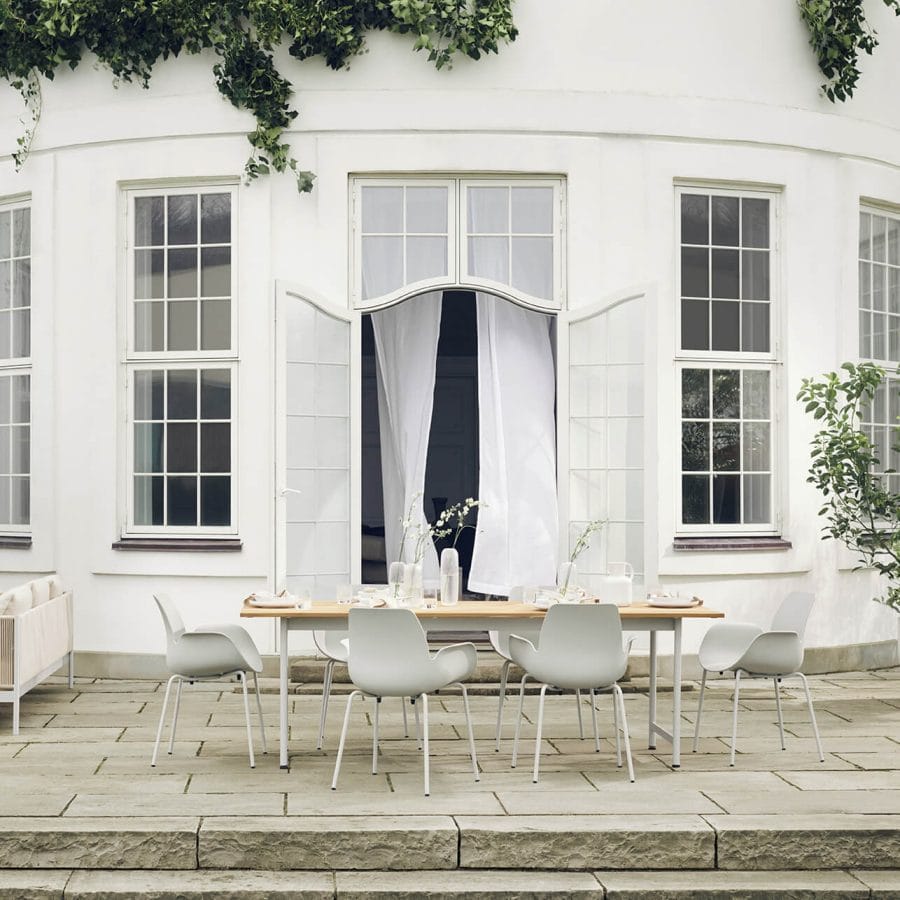 Minimalist outdoor dining table with white chairs set against a classic white-walled residence, elegant draped curtains from a doorway, and delicate vases with fresh flowers on the table.