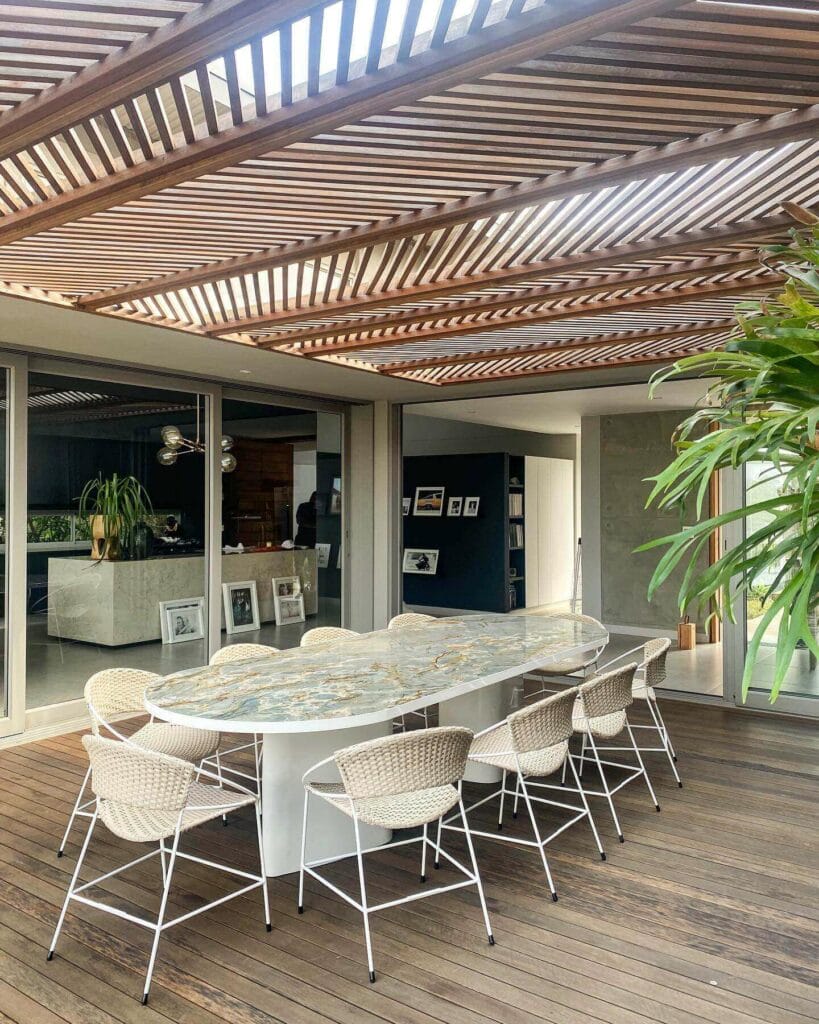 Contemporary outdoor dining area featuring a marbled oval table, wicker chairs with white frames, set on a wooden deck beneath an intricate wooden pergola, adjacent to a modern home interior.