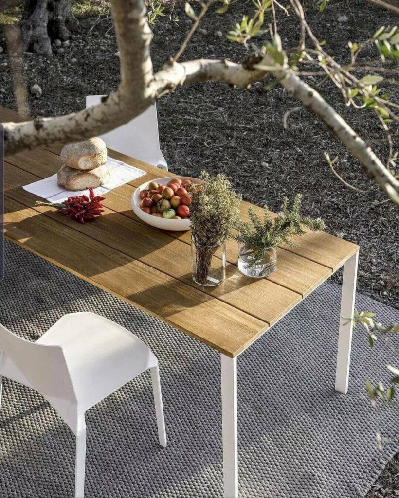 Rustic outdoor dining setup with a wooden slatted table displaying fresh bread, a bowl of assorted fruits, and vases with dried herbs, complemented by a white chair, all set amidst a backdrop of olive trees and woven ground mat.