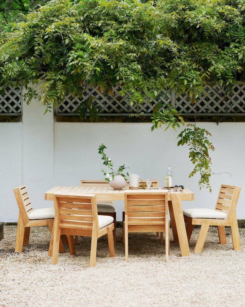 Tranquil outdoor dining space featuring a natural wooden table and chairs set on a pebble ground, backed by a white lattice fence covered in lush green foliage.