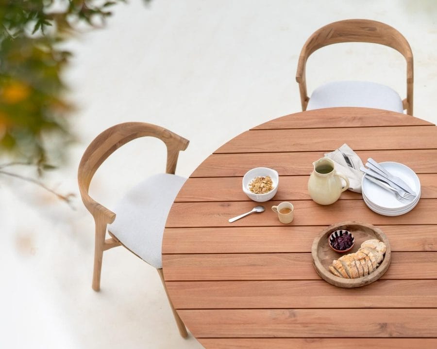 Top-down view of a round wooden outdoor dining table set with breakfast items like a pastry, granola, a pot of tea, and stack of plates, complemented by curved wooden chairs.