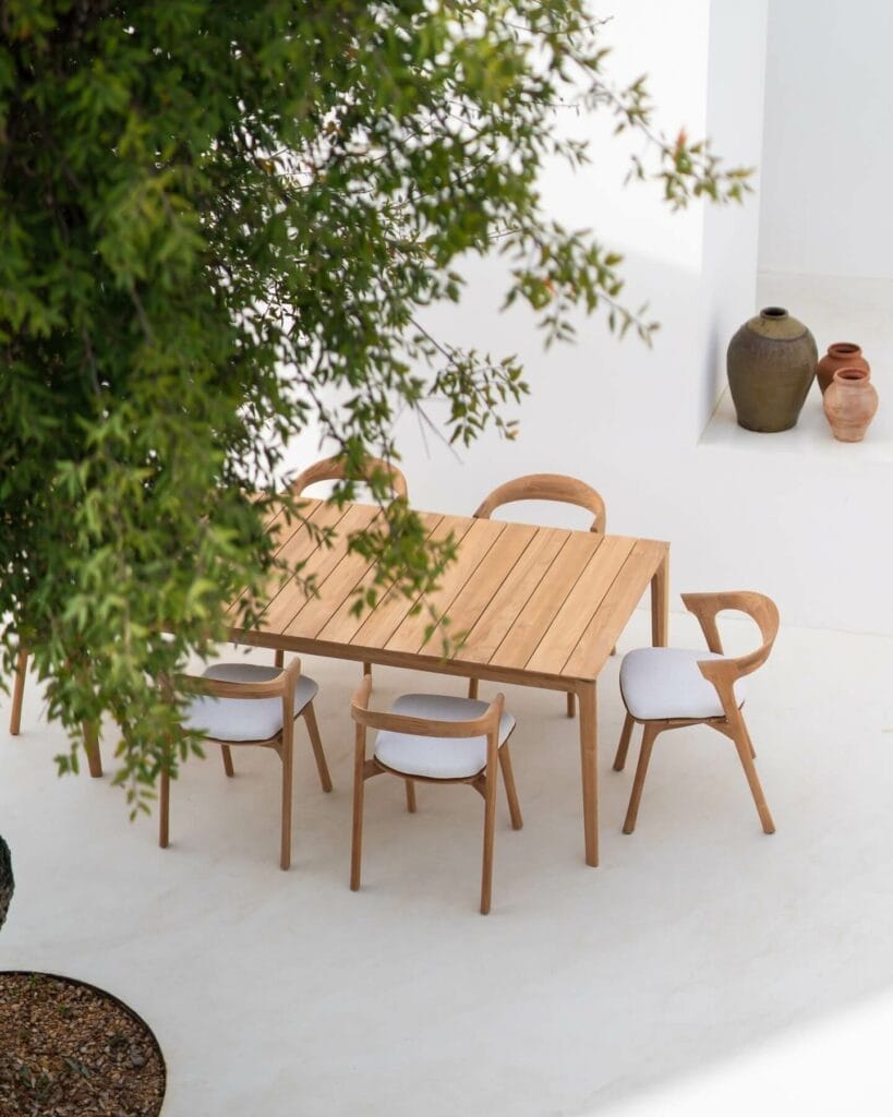 Modern wooden outdoor dining table accompanied by matching curved-back chairs, set against a serene backdrop with overhanging greenery and decorative clay pots.