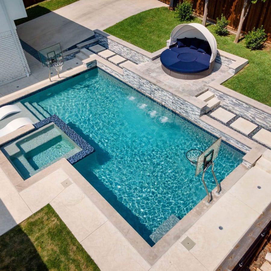 pool with a built-in square hot tub and waterfall, flanked by stone loungers and a round shaded daybed on a beige deck.