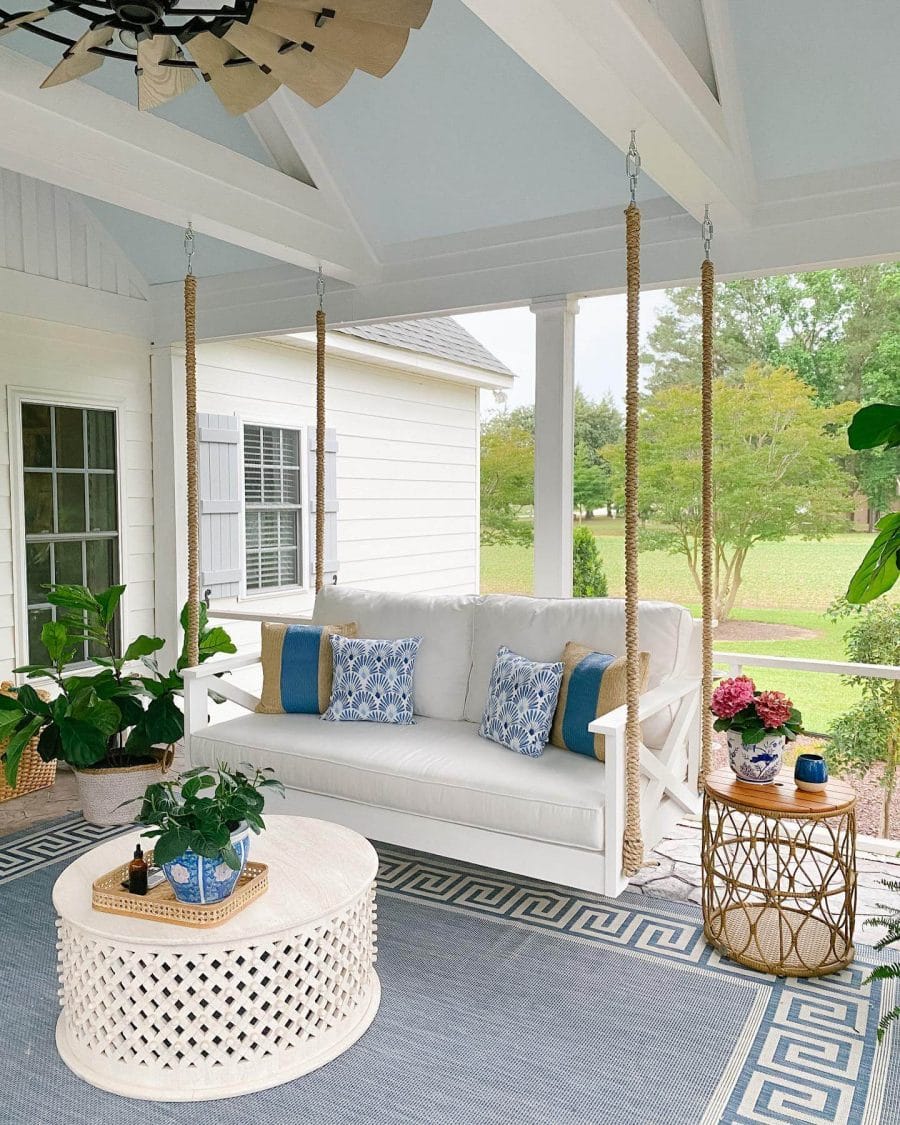 Outdoor living space with a hanging swing sofa in whitewashed wood, blue floral cushions, and a round lattice coffee table. Surrounded by greenery.