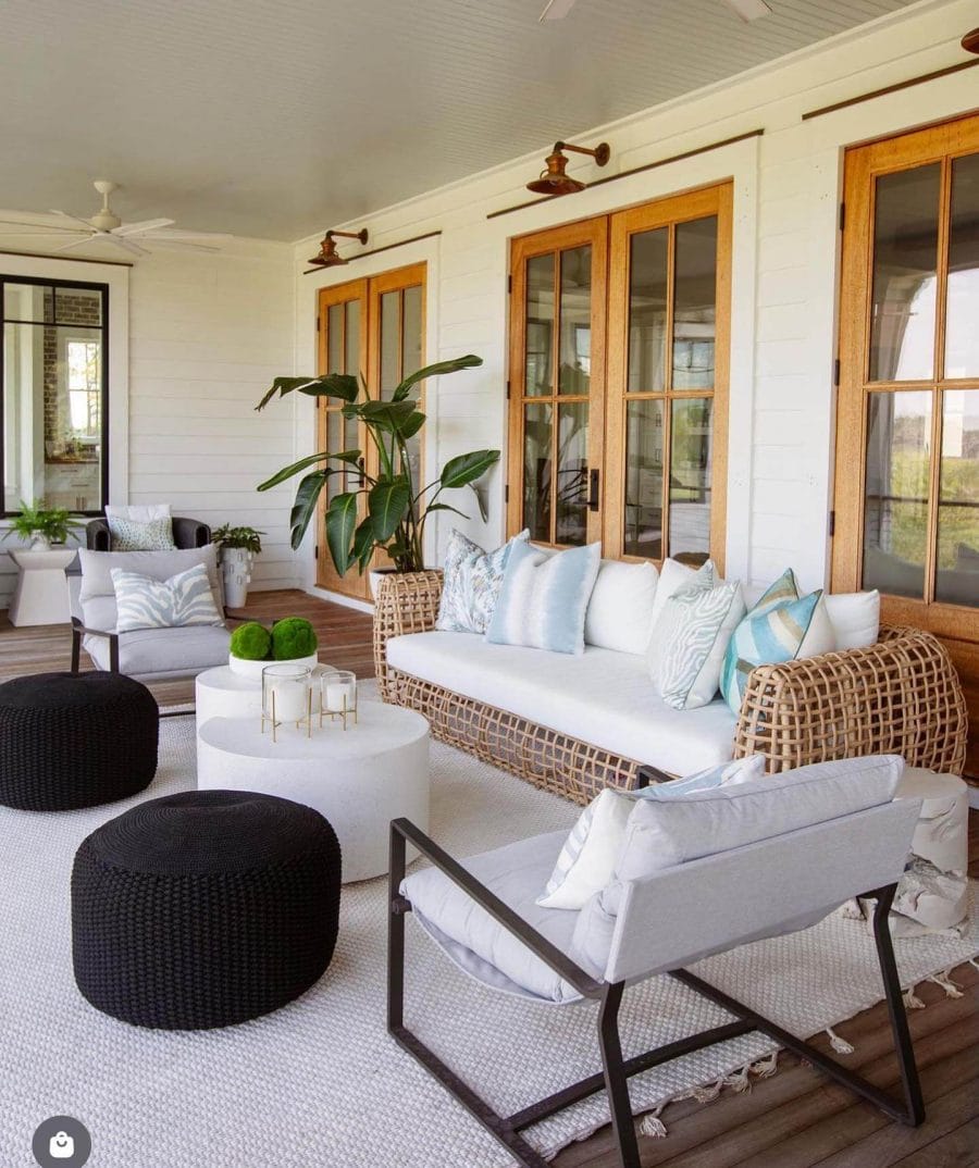 Spacious back porch with a woven rattan sofa, white cushions, and a circular coffee table, surrounded by lush plants, creating a cozy outdoor living space.