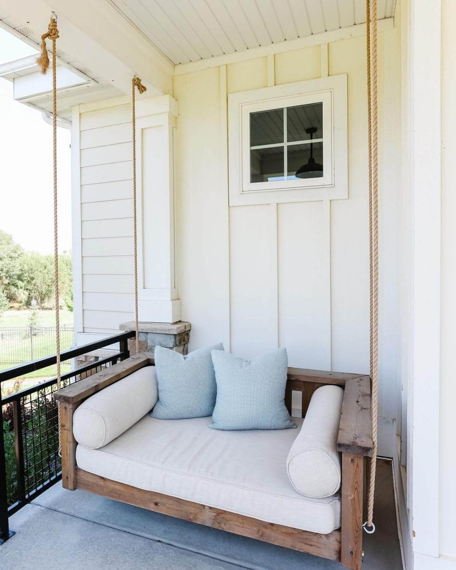 Cozy back porch featuring a wooden swing with beige cushions, surrounded by a black metal railing and protected by a roof overhang.
