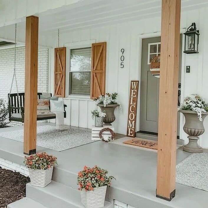 Cozy back porch featuring wooden posts, a swinging bench, potted flowers, a welcome sign, and a modern farmhouse design.