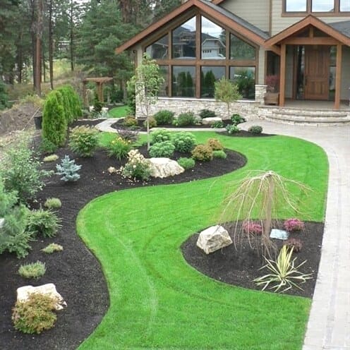 Manicured lawn with a curving pathway leads to a modern home, accented by shrubs, a weeping willow, and black mulch beds.