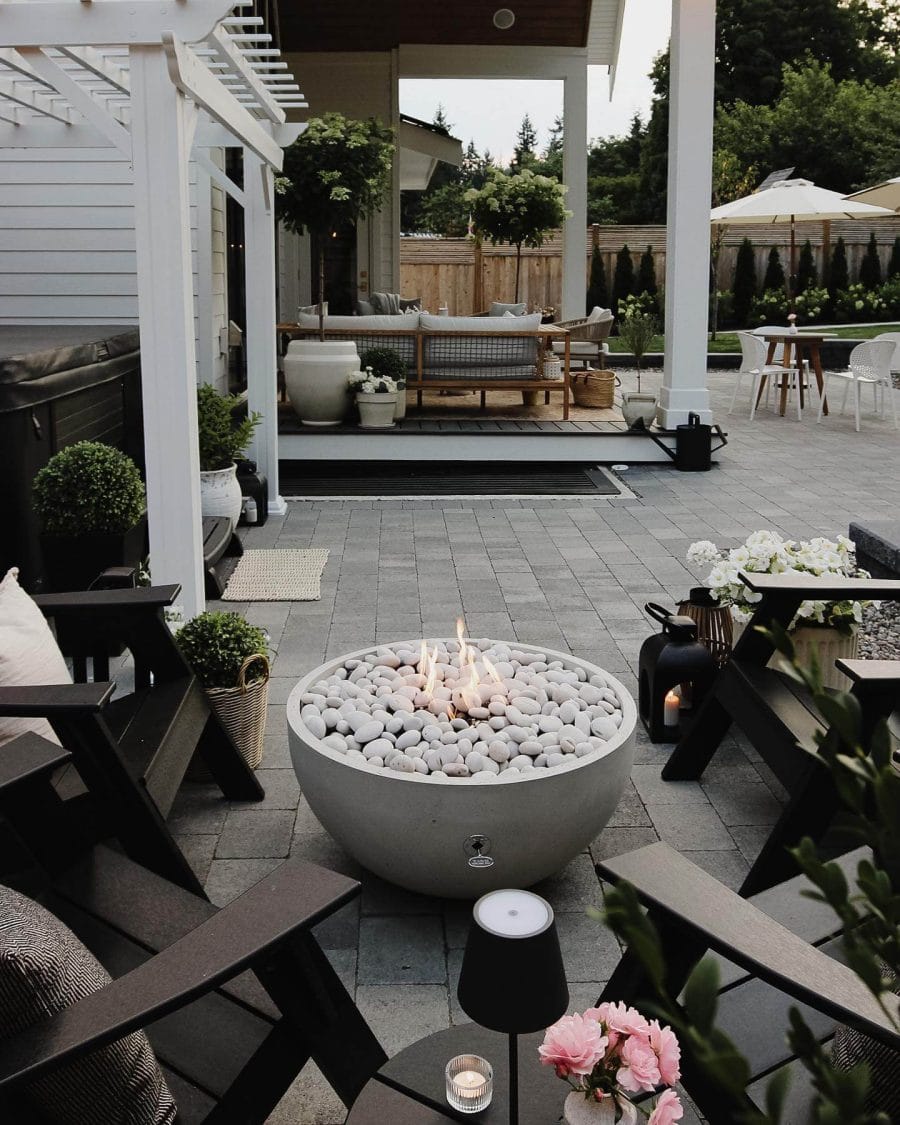Outdoor living space with a circular concrete fire pit surrounded by black wooden chairs, paving stones, and potted plants for a cozy, budget-friendly design.
