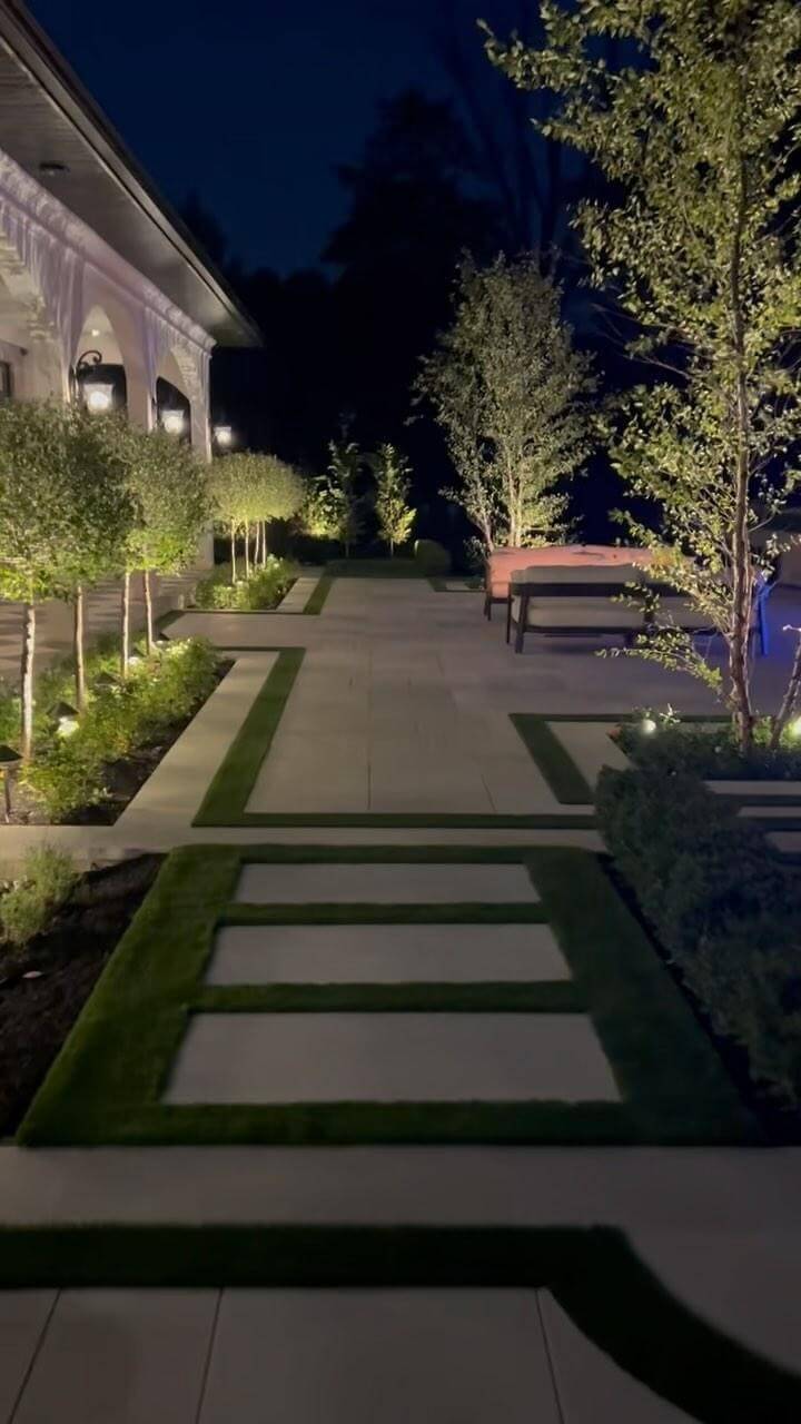Modern backyard landscaping features clean lines with concrete pathways and artificial grass, surrounded by low shrubs and ornamental trees. A bench enhances social interaction.