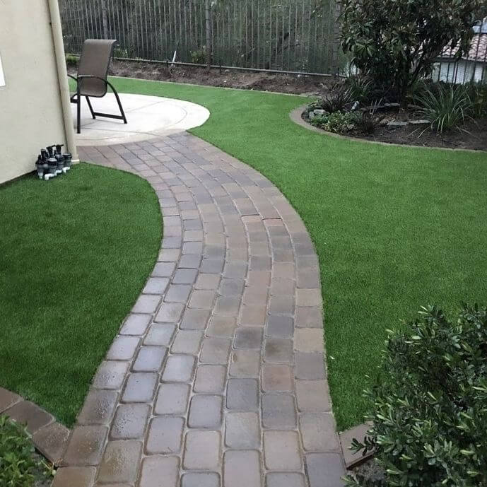 Curved pathway of interlocking pavers leads to a circular seating area, surrounded by low-maintenance artificial turf and a small hedge.