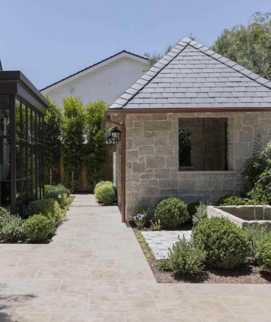 Garden landscape features a stone pathway linking a rustic stone building and a modern glass structure, surrounded by boxwood and lavender.