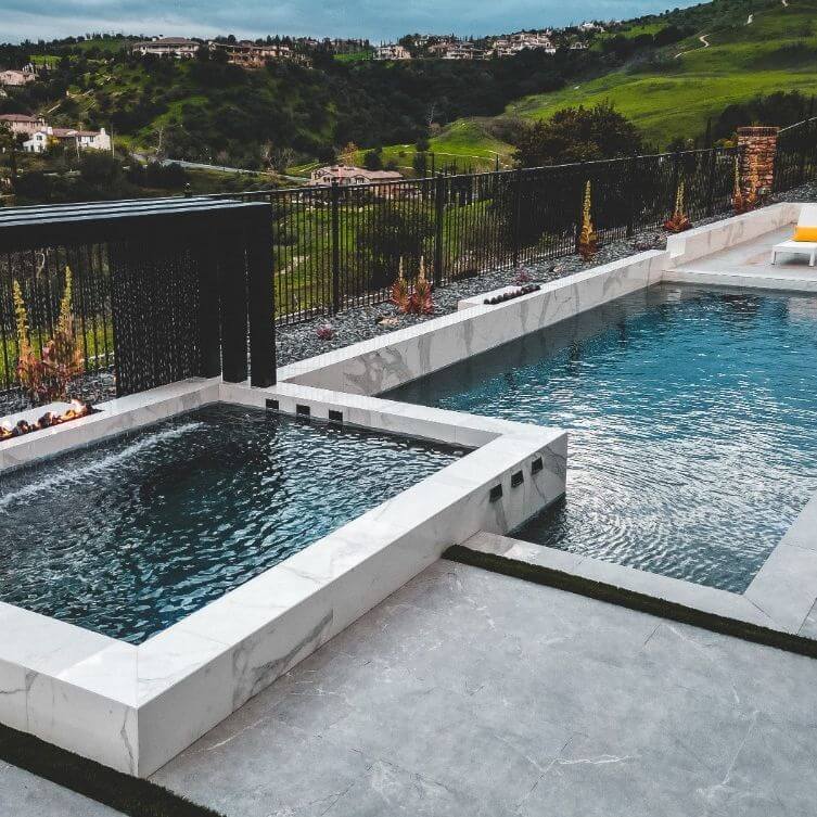 Luxurious backyard featuring two white marble pools, a serene water feature, and lush greenery, bordered by elegant black fencing.
