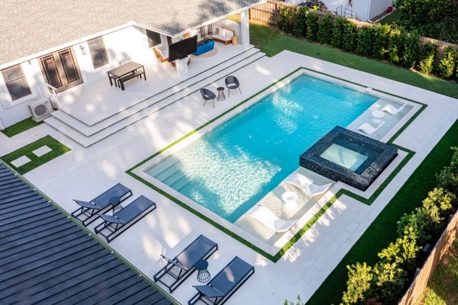 Luxurious backyard with a rectangular swimming pool, dark stone hot tub, and elegant stone tiles, surrounded by lush green grass.