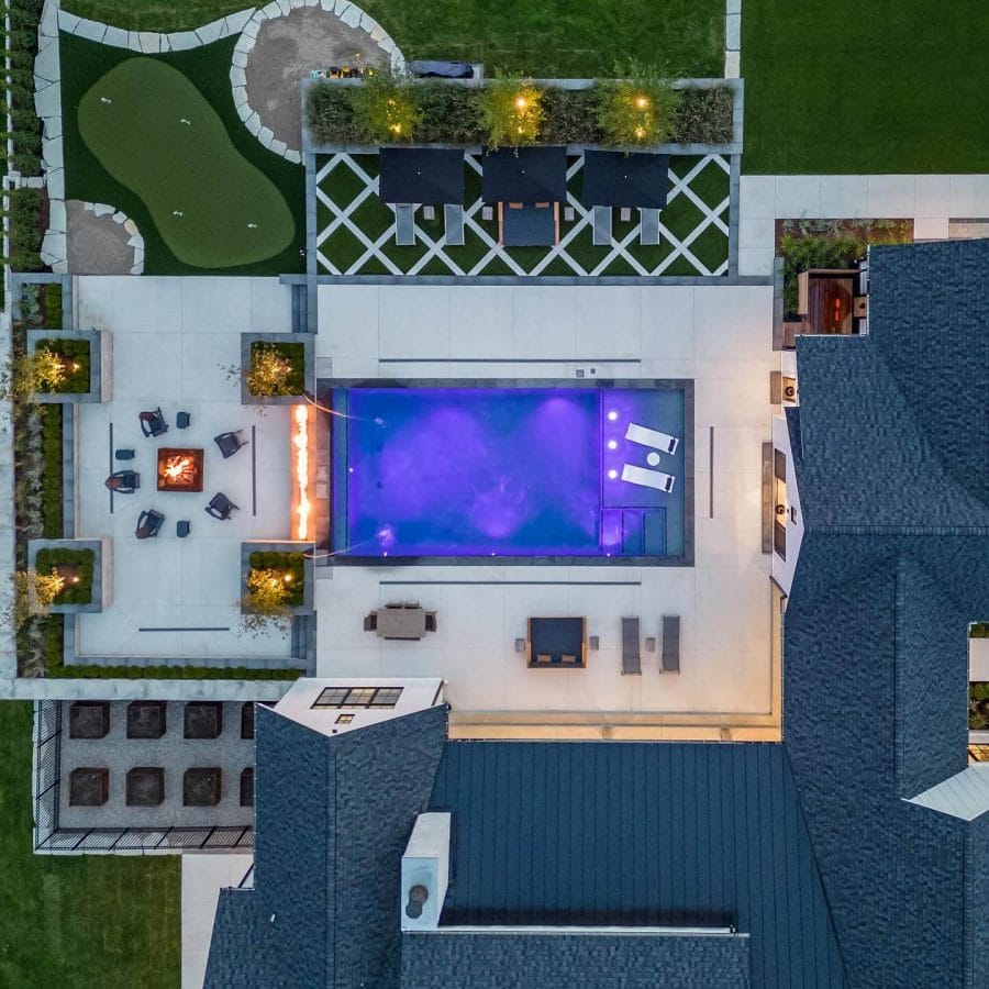 Expansive backyard oasis with a rectangular pool illuminated by purple lights, sleek concrete decking, cozy fire pit, and lush greenery.