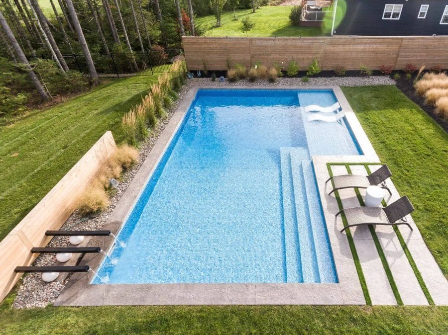 Crystal-clear rectangular swimming pool in a lush backyard, framed by lounge chairs, vibrant plants, and a modern wooden fence.