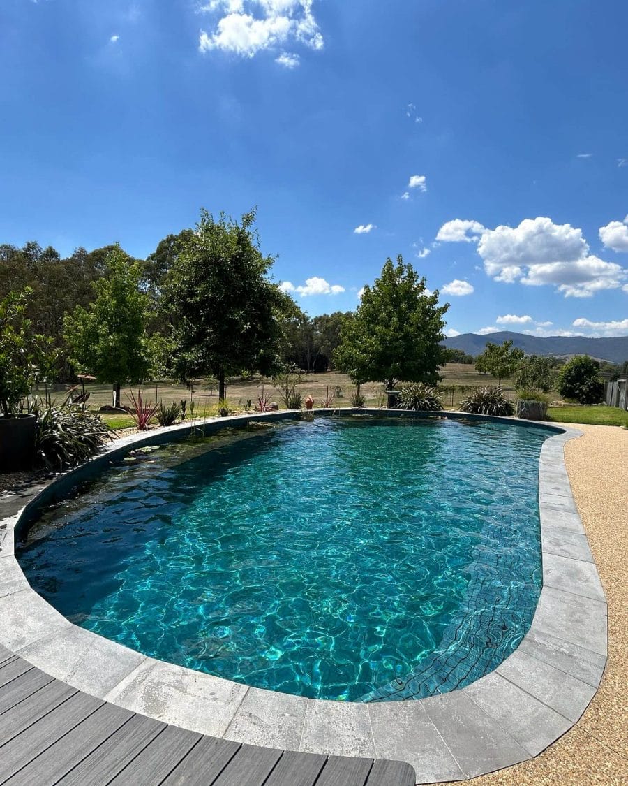 A beautifully designed custom pool surrounded by lush greenery, with clear blue water, framed by gray stone coping and a wooden deck.