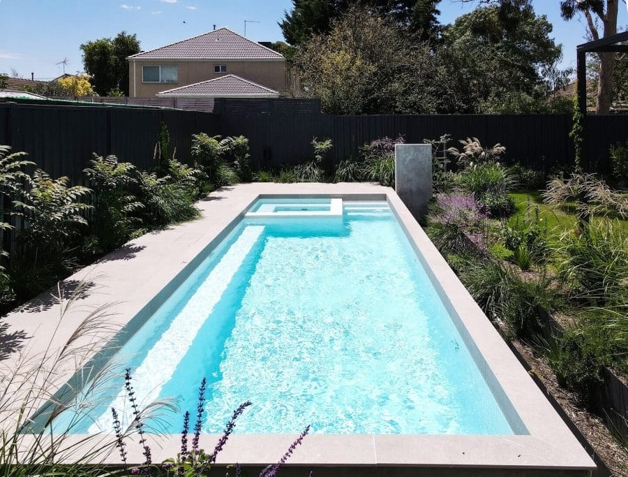 Luxurious rectangular swimming pool with crystal-clear water, bordered by light gray concrete, surrounded by lush greenery and vibrant purple flowers.