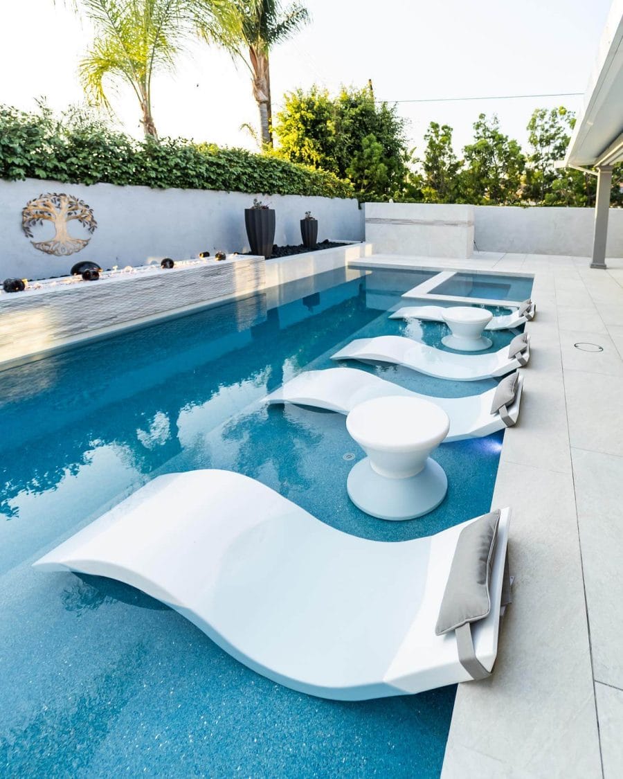 Luxurious custom pool area featuring sleek white lounge chairs and a minimalist side table, surrounded by lush greenery and artistic decor.