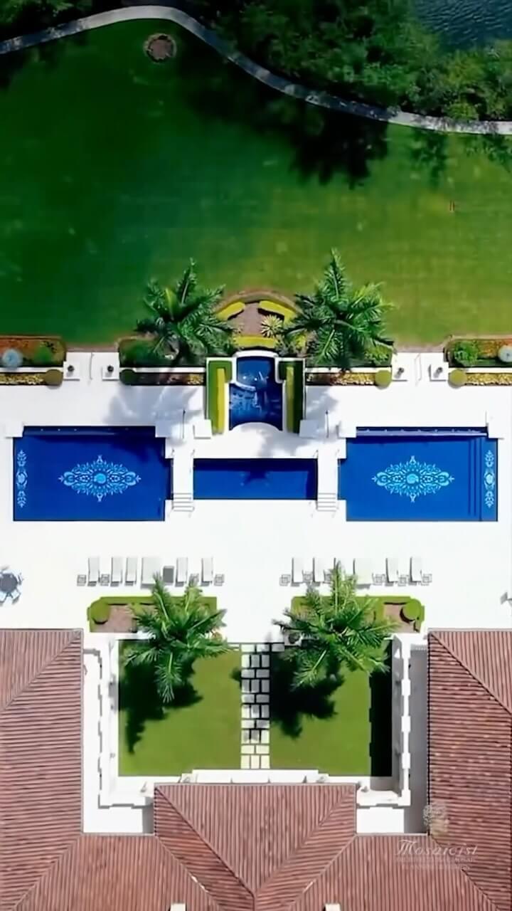 Aerial view of a tropical resort-style backyard featuring two deep blue pools, a central waterfall, and palm trees, surrounded by lounge chairs and terracotta pavilions.