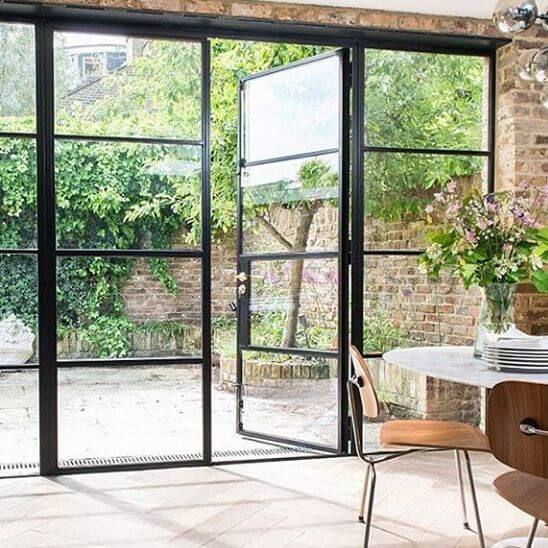 Sleek black steel-framed glass doors open to a walled courtyard, showcasing climbing ivy, a pale stone patio, and a cozy dining nook.