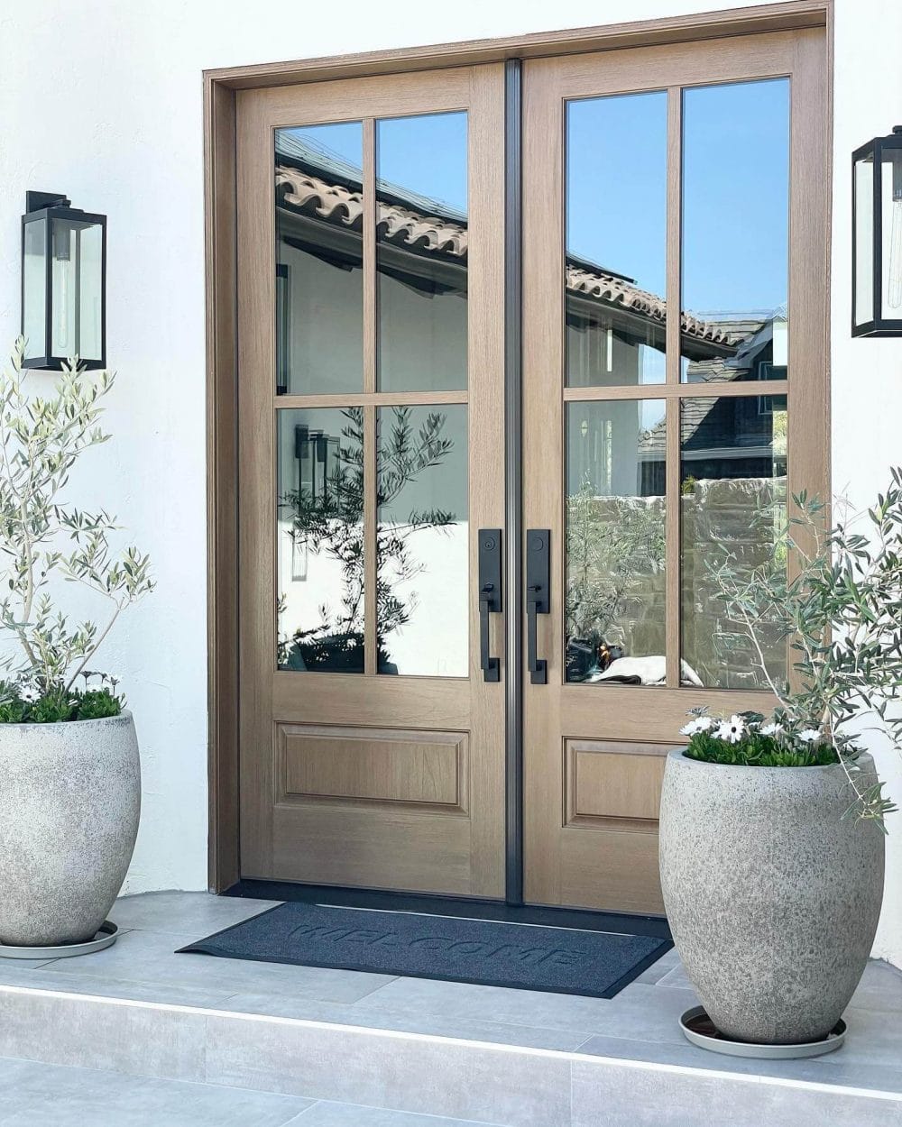 Twin light-oak doors with glass panes frame a serene Mediterranean entry, flanked by stone planters with dwarf olive trees and seasonal flowers.