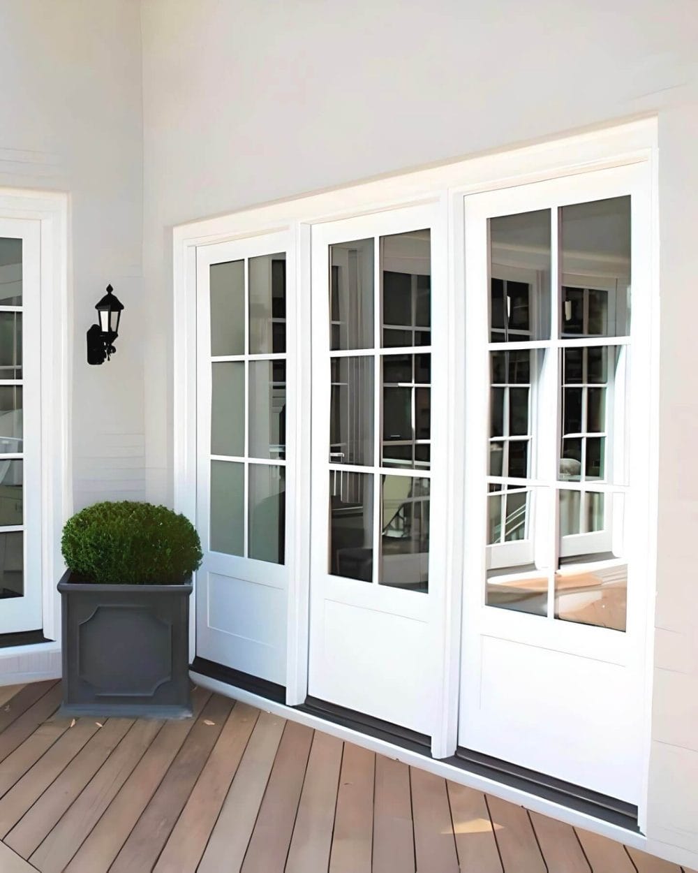 Bright backyard porch features triple white French doors leading to a tan diagonal deck, accented by a charcoal planter with boxwood.